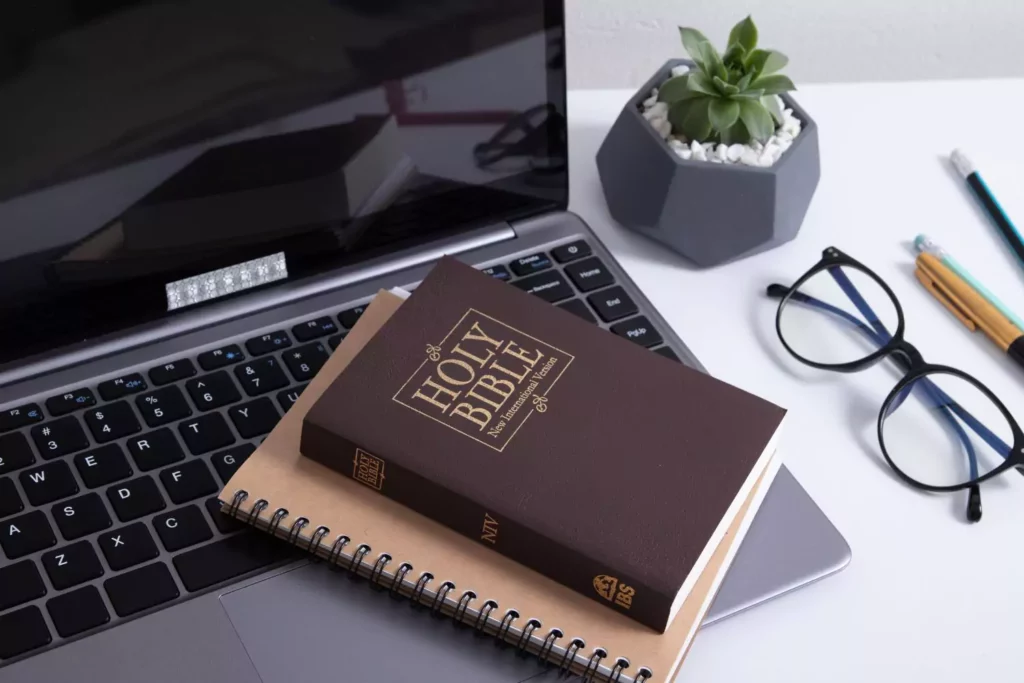 Desk with a bible and notebook on top of a laptop with a plant and glasses next to the laptop.