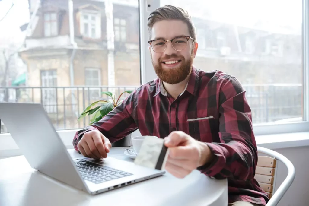 Guy in front of laptop looking at the camera extending his credit card toward the camera. Spending money.