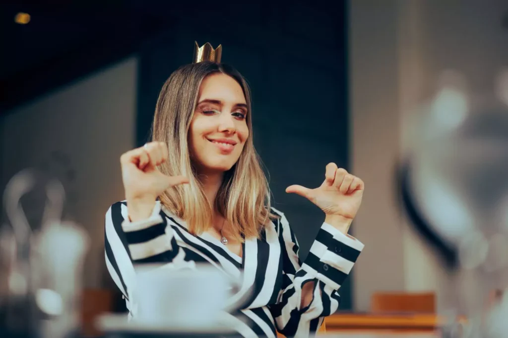 a woman with a crown on her head, pointing her thumbs at herself. She's self-righteous