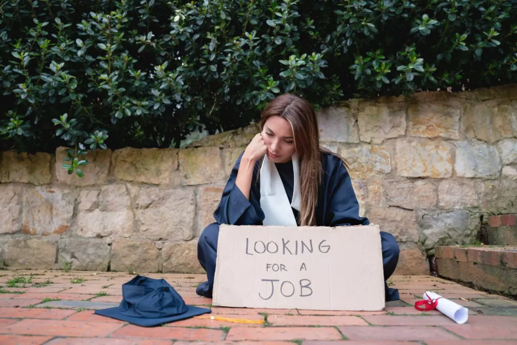 a woman in a graduation gown without a job, sitting on the ground, with a cardboard sign that says "looking for a job"
