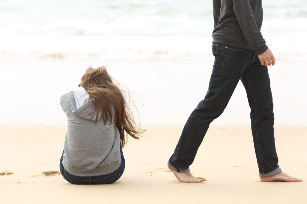 man walking away from girl, who's sitting, on a beach
