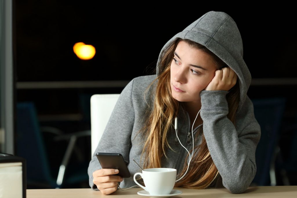 young woman sitting in coffeeshop, holding phone, looking sadly outside window.