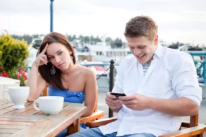 woman and man on a date, woman is frustrated, man is laughing at something on his phone, she's thinking Why am I attracted to this guy?