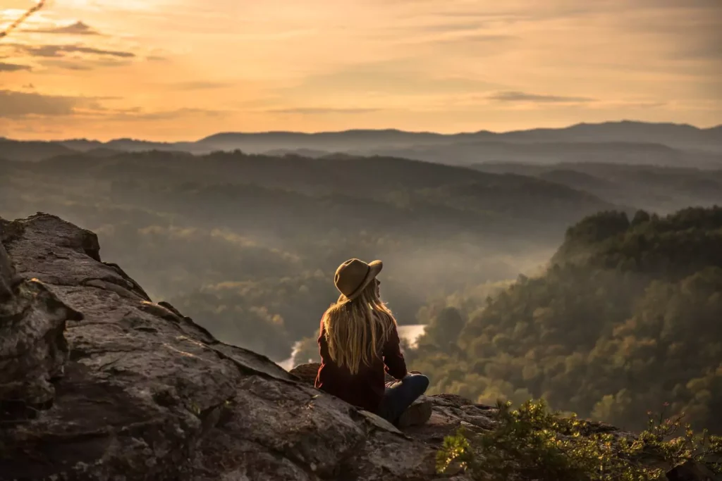 a woman sitting on a mountain looking out over the landscape at dusk pondering humility