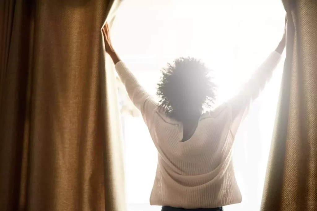 Woman opening the curtains with the sun shining through. Trust God with all things.