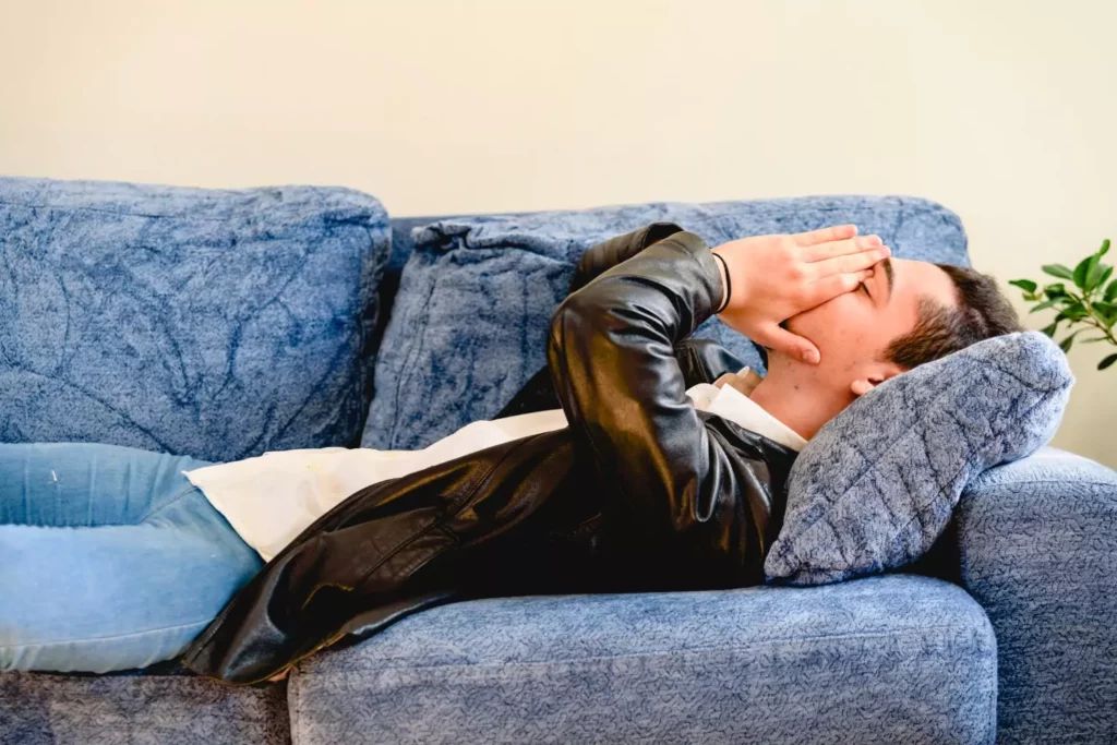 young man lying on a blue couch with his hands over his face thinking about new changes in his life