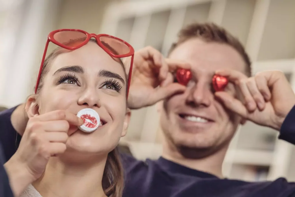 woman with valentine's glasses on her head, holding a coin with lips on it in front of her mouth, a man holding heart shaped candies in front of his eyes, creative date ideas