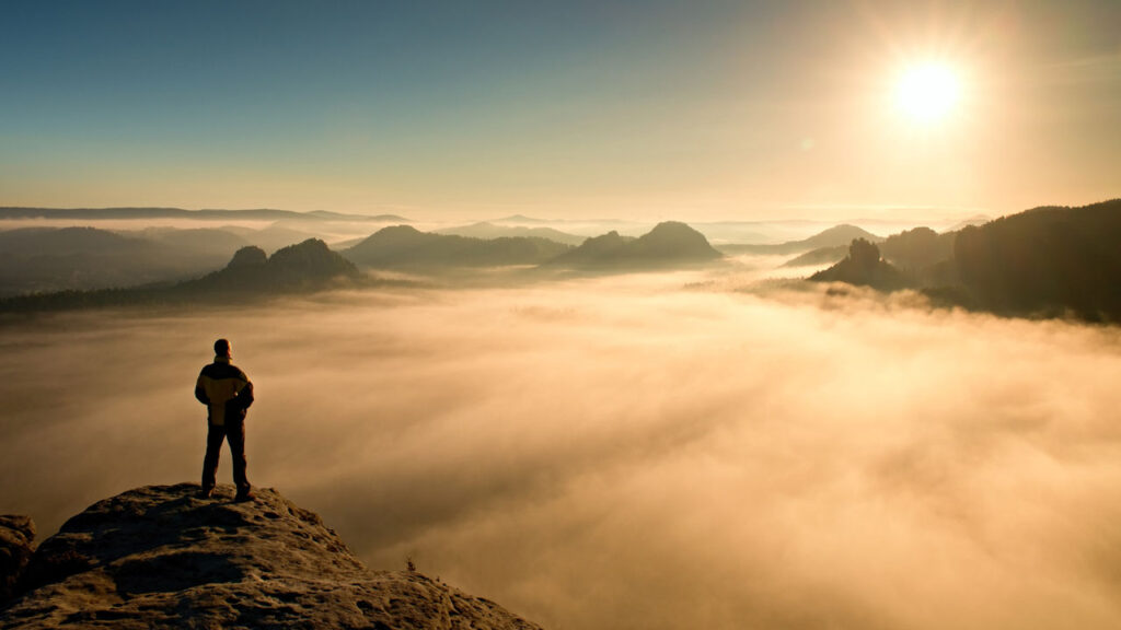 Man standing on a high mountain top above the clouds, looking toward the sun