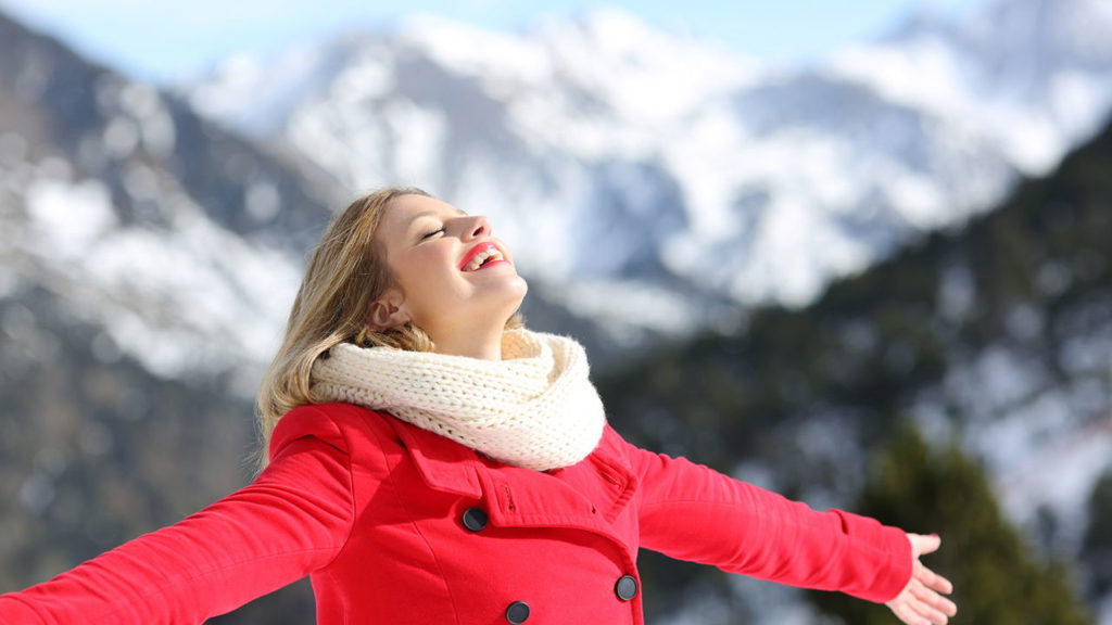 woman, arms outstretched, wearing a red jacket, standing in front of snowy mountains